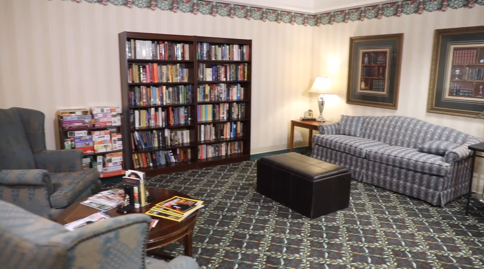 A cozy living room area with a patterned carpet, a striped sofa, two upholstered armchairs, a wooden coffee table with magazines, a black ottoman, a wooden bookshelf filled with books, a side table with a lamp, and framed pictures on the wall.