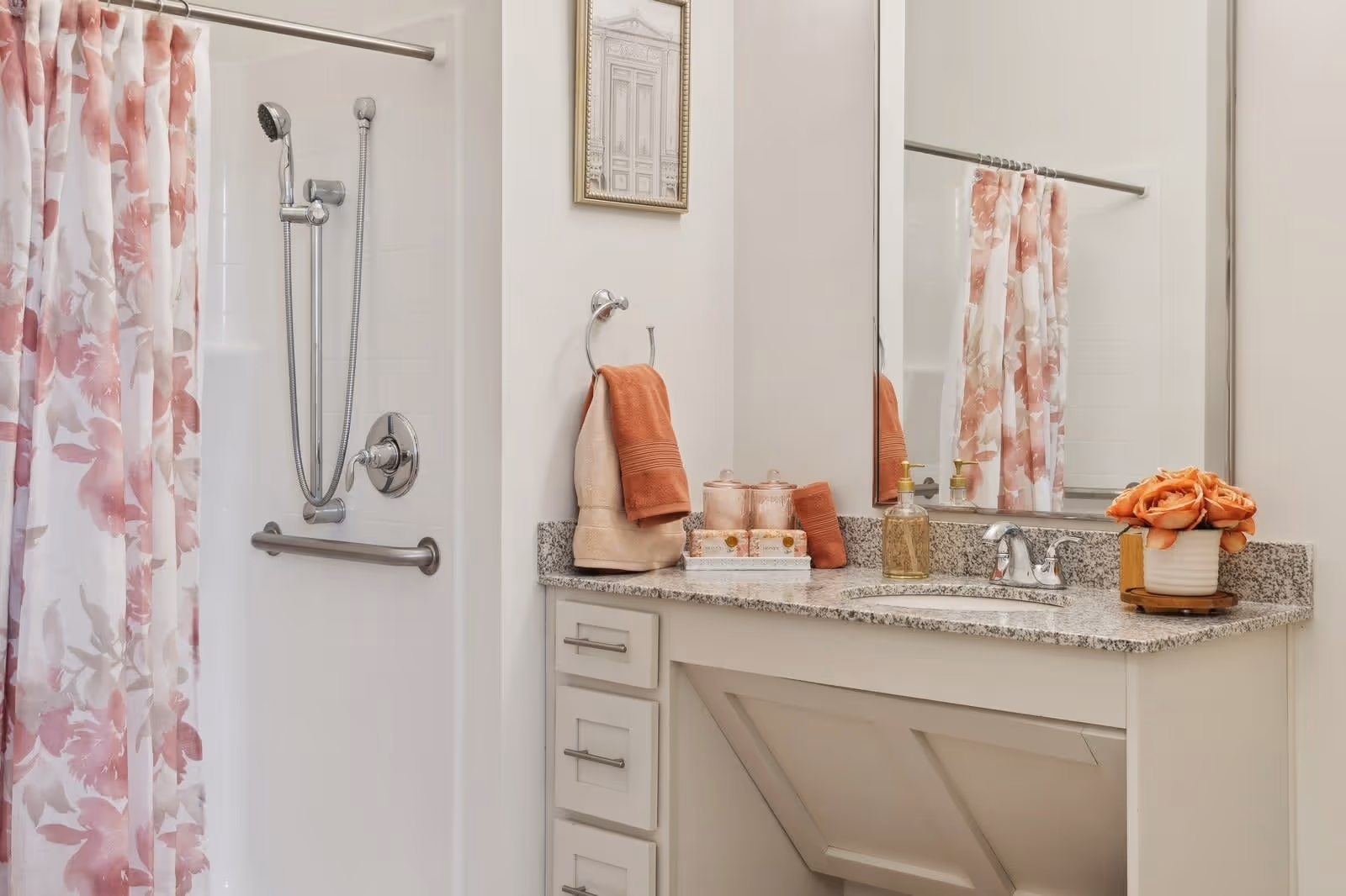 A bathroom with a shower area featuring a floral pink and white shower curtain, a handheld showerhead, and a grab bar. Next to the shower is a vanity with a granite countertop, a sink, a large mirror, and decorative items including towels, soap dispensers, and a small vase with orange flowers.