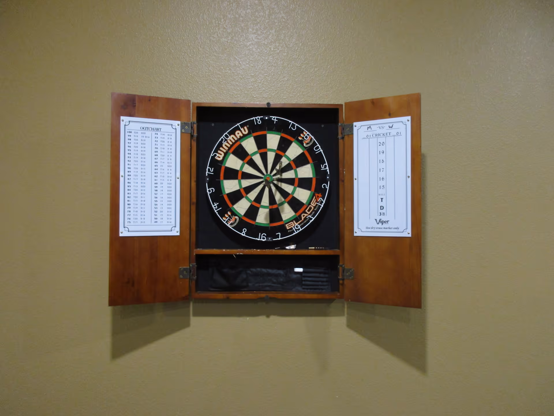 A dartboard mounted on a beige wall inside a wooden cabinet with open doors. The cabinet doors have printed charts attached to them, one showing an outchart and the other a cricket score sheet. The dartboard is a Winmau Blade 4 model.