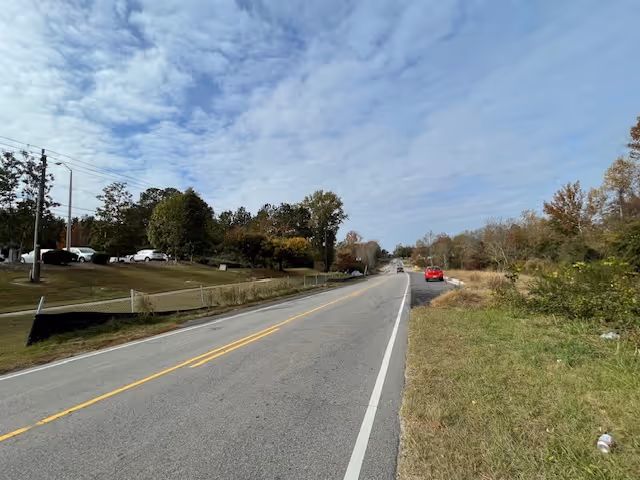 A two-lane road with a yellow dividing line stretches into the distance under a partly cloudy sky. On the left side of the road, there is a grassy embankment with several parked cars and trees behind a fence. On the right side, there is a grassy area with some scattered bushes and trees further back. A red car is parked on the right shoulder of the road.