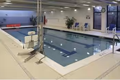Indoor swimming pool area with a pool lift for accessibility, lane markings in the water, several chairs along the far wall, and large windows letting in natural light.