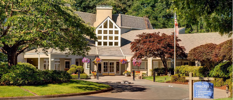 Front exterior view of SpringRidge At Charbonneau facility showing a well-maintained building with a peaked roof, large windows, hanging flower baskets, an American flag, and landscaped greenery including trees and bushes.