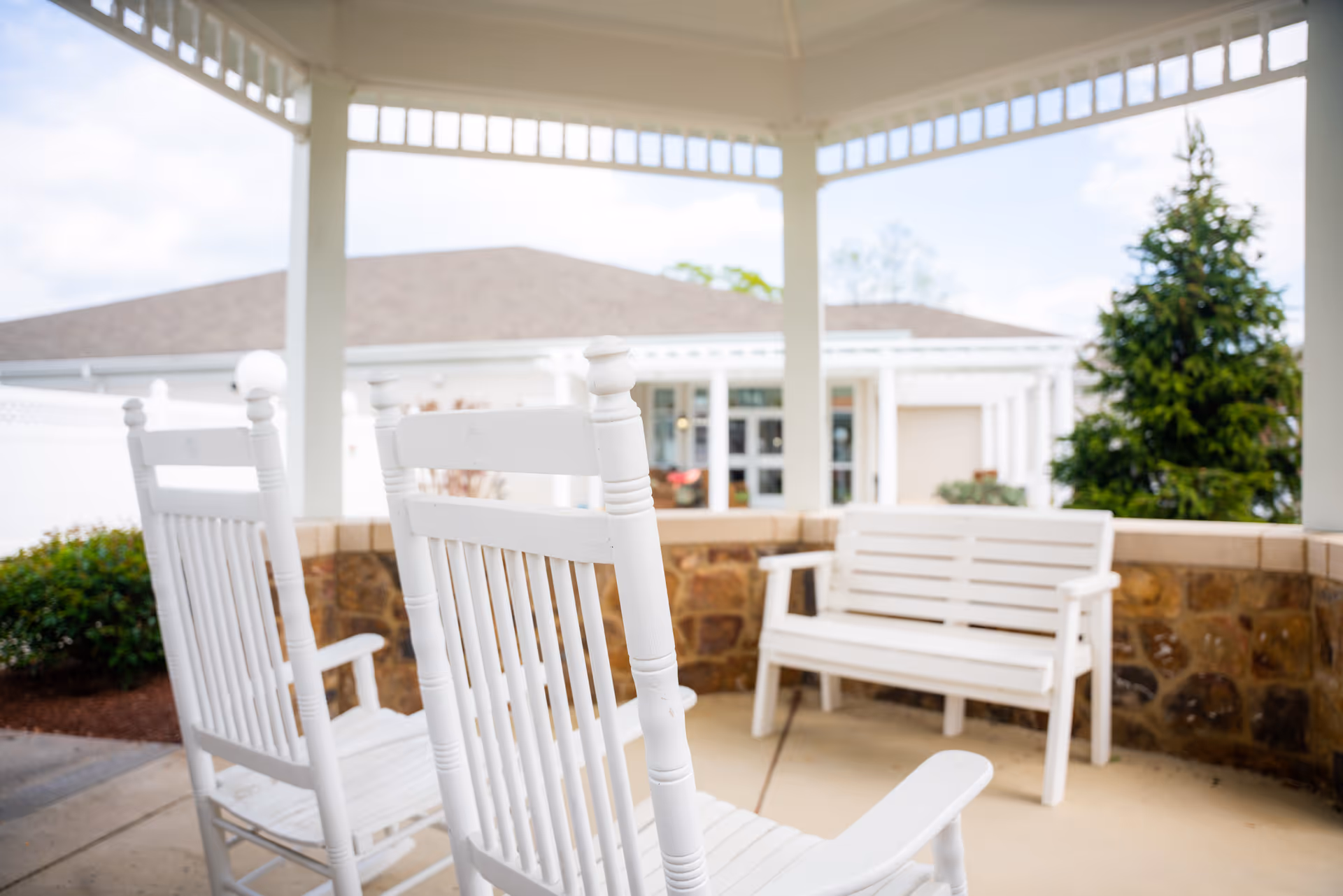 White wooden rocking chairs and a white wooden bench under a covered outdoor patio area with a stone half-wall, with a building and greenery visible in the background.