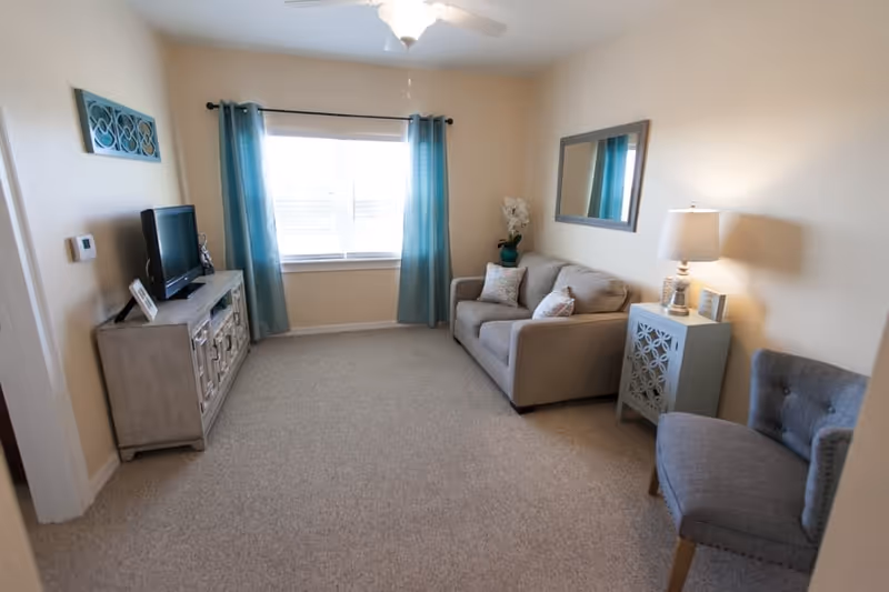 A cozy living room with beige walls and carpeted floor. The room features a small beige sofa with decorative pillows, a gray upholstered chair, a side table with a lamp, a wall mirror, and a TV on a wooden cabinet. Blue curtains frame a window letting in natural light.