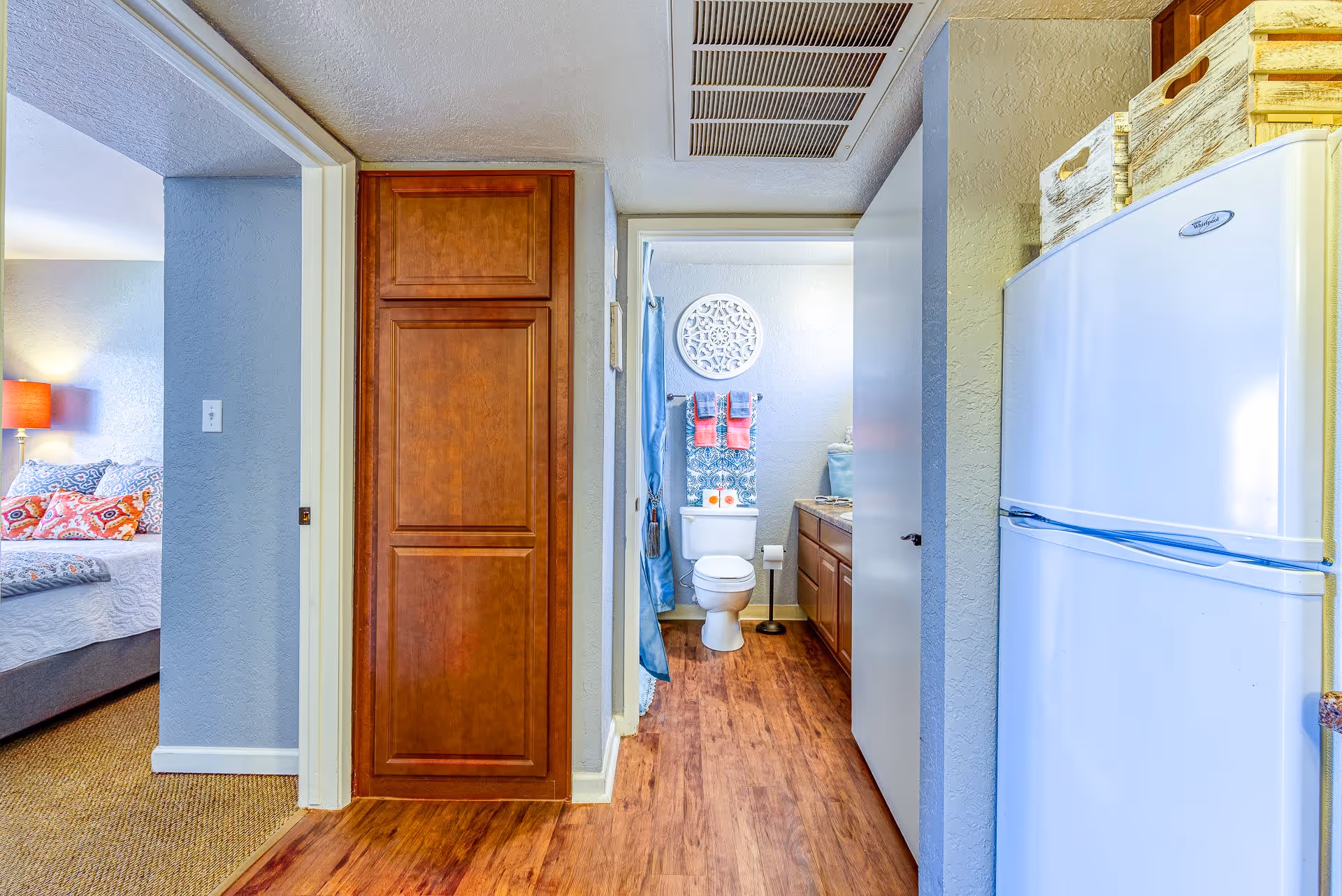 View down a hallway showing a bathroom with a toilet, sink, and shower curtain. To the left is a bedroom with a bed, pillows, and a lamp. To the right is a white refrigerator with baskets on top. The floor is wood and the walls are light blue.