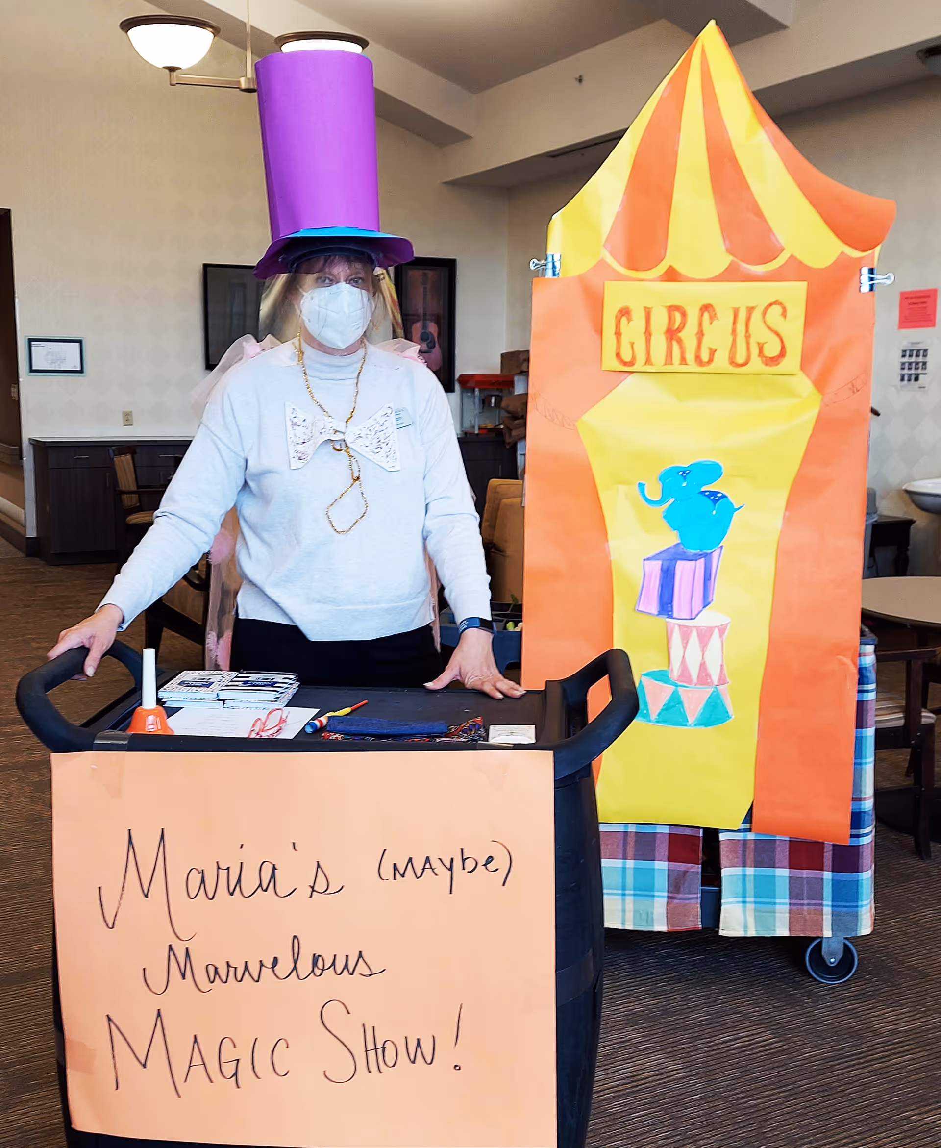 A person wearing a purple tall hat, a face mask, and a large bow tie stands behind a cart with a sign that reads 'Maria's (maybe) Marvelous MAGIC SHOW!'. Behind them is a colorful circus-themed backdrop with an illustration of a blue elephant balancing on circus pedestals. The setting appears to be an indoor common area with tables and chairs.