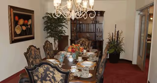 A formal dining room with an ornate wooden dining table set with china, glassware, and a floral centerpiece. The room features upholstered chairs with intricate patterns, a chandelier hanging above the table, a large wooden cabinet against the back wall, a potted plant in the corner, and a framed floral painting on the wall.