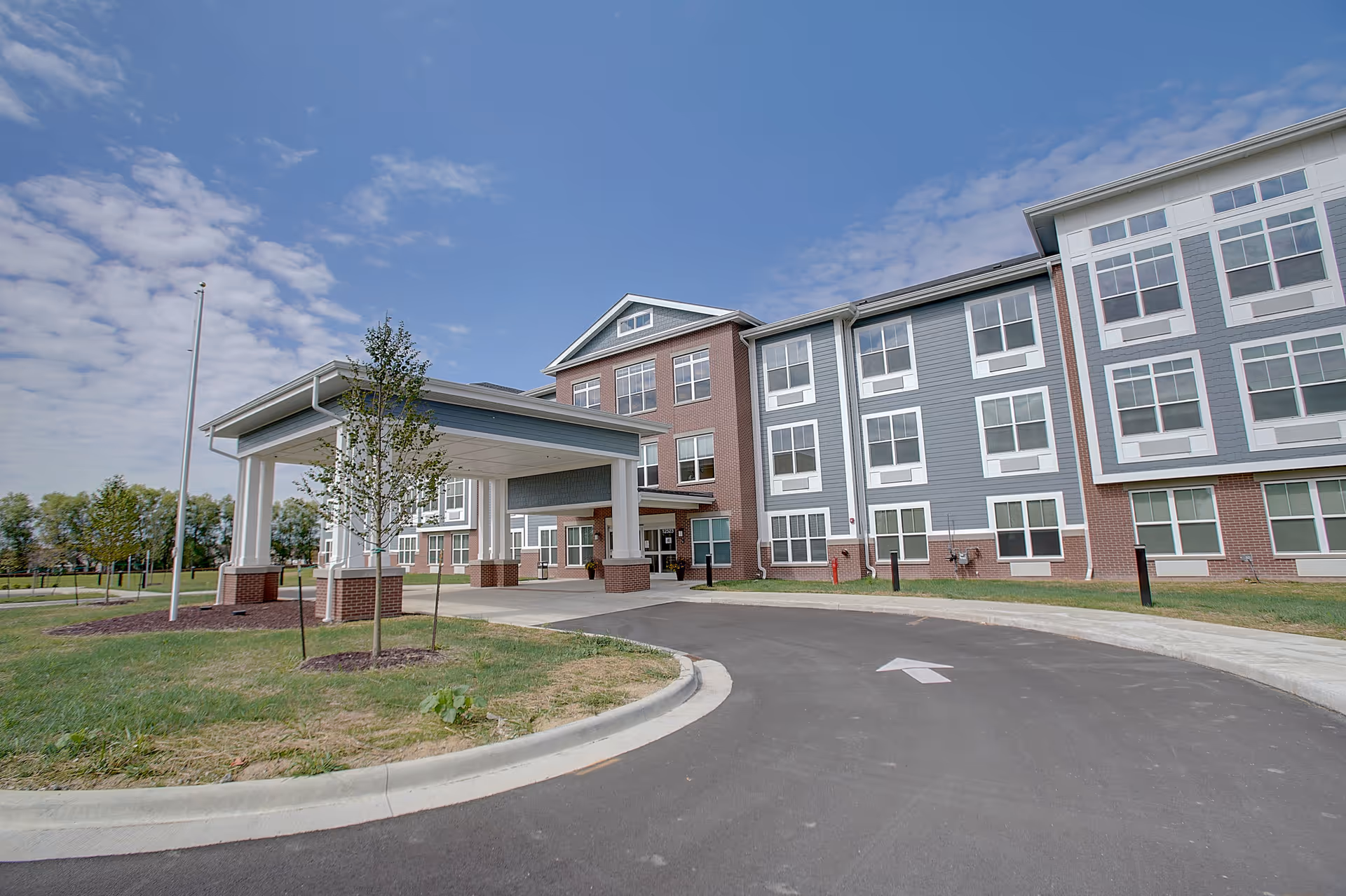 Exterior view of a modern three-story senior living facility with a covered entrance driveway, surrounded by a lawn and young trees under a partly cloudy sky.