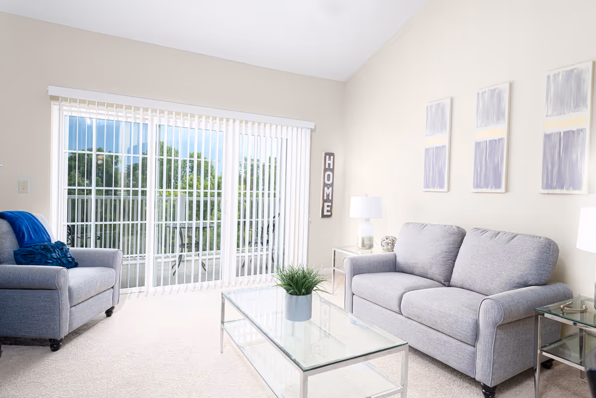 A bright living room with a gray sofa and matching armchair, a glass coffee table with a small green plant, two side tables with lamps, three abstract paintings on the wall, and large sliding glass doors with vertical blinds leading to a balcony with outdoor furniture and greenery visible outside.