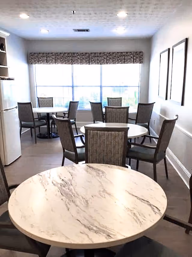 A bright dining area with several round tables and cushioned chairs arranged around them. Large windows with patterned valances allow natural light to fill the room. The walls are light-colored with framed artwork, and a refrigerator is visible on the left side.