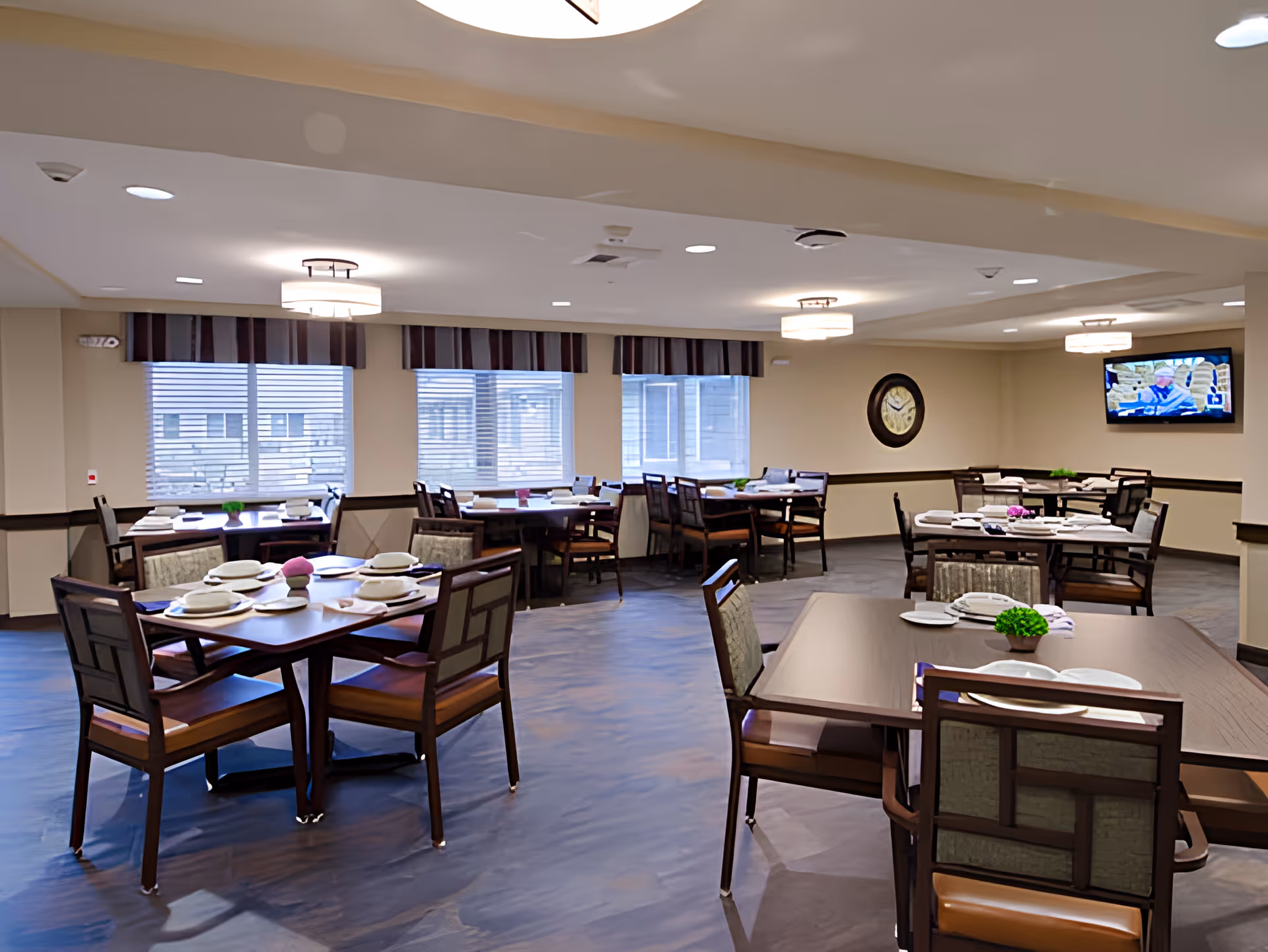 A dining room in a senior living facility with multiple wooden tables and chairs arranged neatly. Each table is set with plates, napkins, and small decorative plants. The room has large windows with blinds, ceiling lights, a wall clock, and a television mounted on the wall.