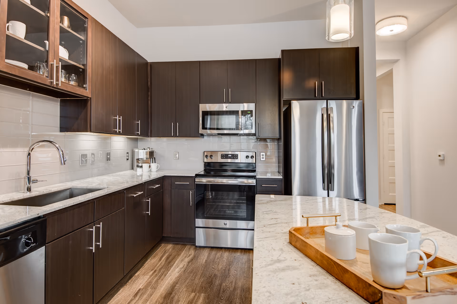 Modern kitchen with dark wood cabinets, stainless steel appliances including a refrigerator, oven, microwave, and dishwasher. White marble countertops with a wooden tray holding white mugs and a sugar bowl. The kitchen has a light-colored tile backsplash and wood flooring.
