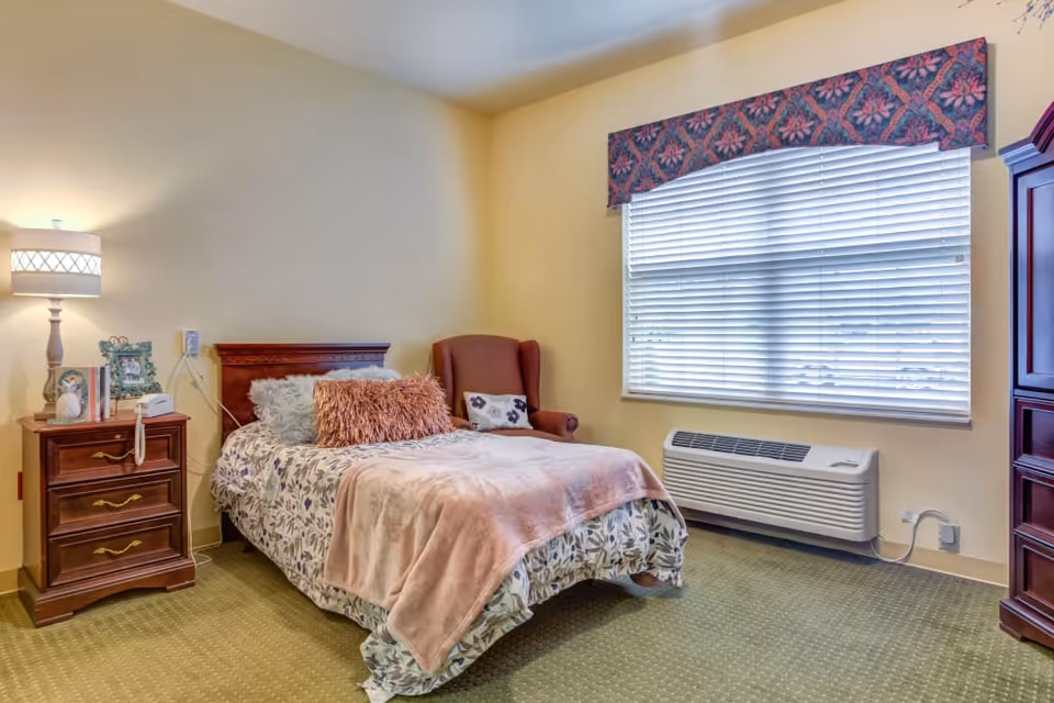 A cozy single bedroom with a bed, bedside table and lamp, an upholstered chair, a large window with blinds, and an HVAC unit.