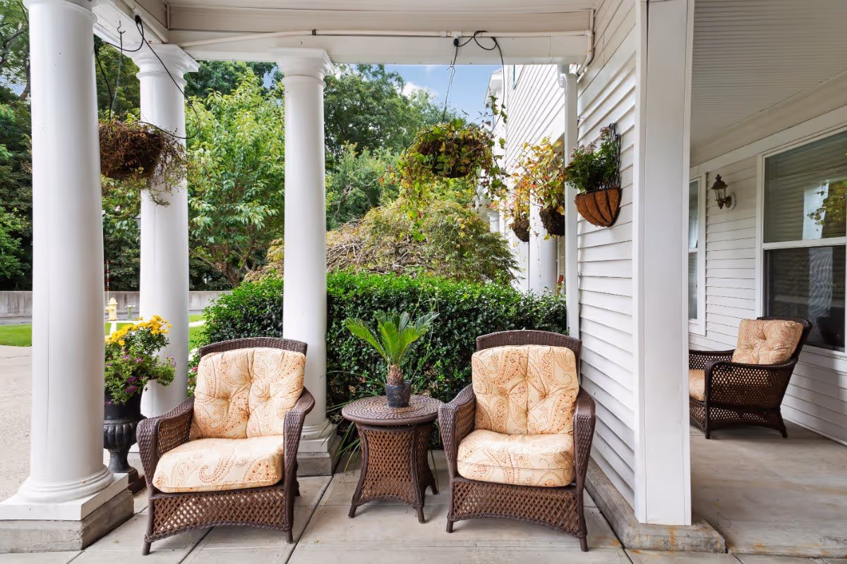 A covered outdoor patio area with two cushioned wicker chairs and a small wicker table with a potted plant between them. The patio is supported by white columns and has hanging flower baskets. There is greenery and trees visible in the background.