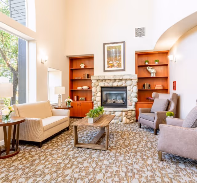 Bright senior living lounge with a stone fireplace flanked by built-in shelves, sofas and armchairs around a wooden coffee table near large windows.