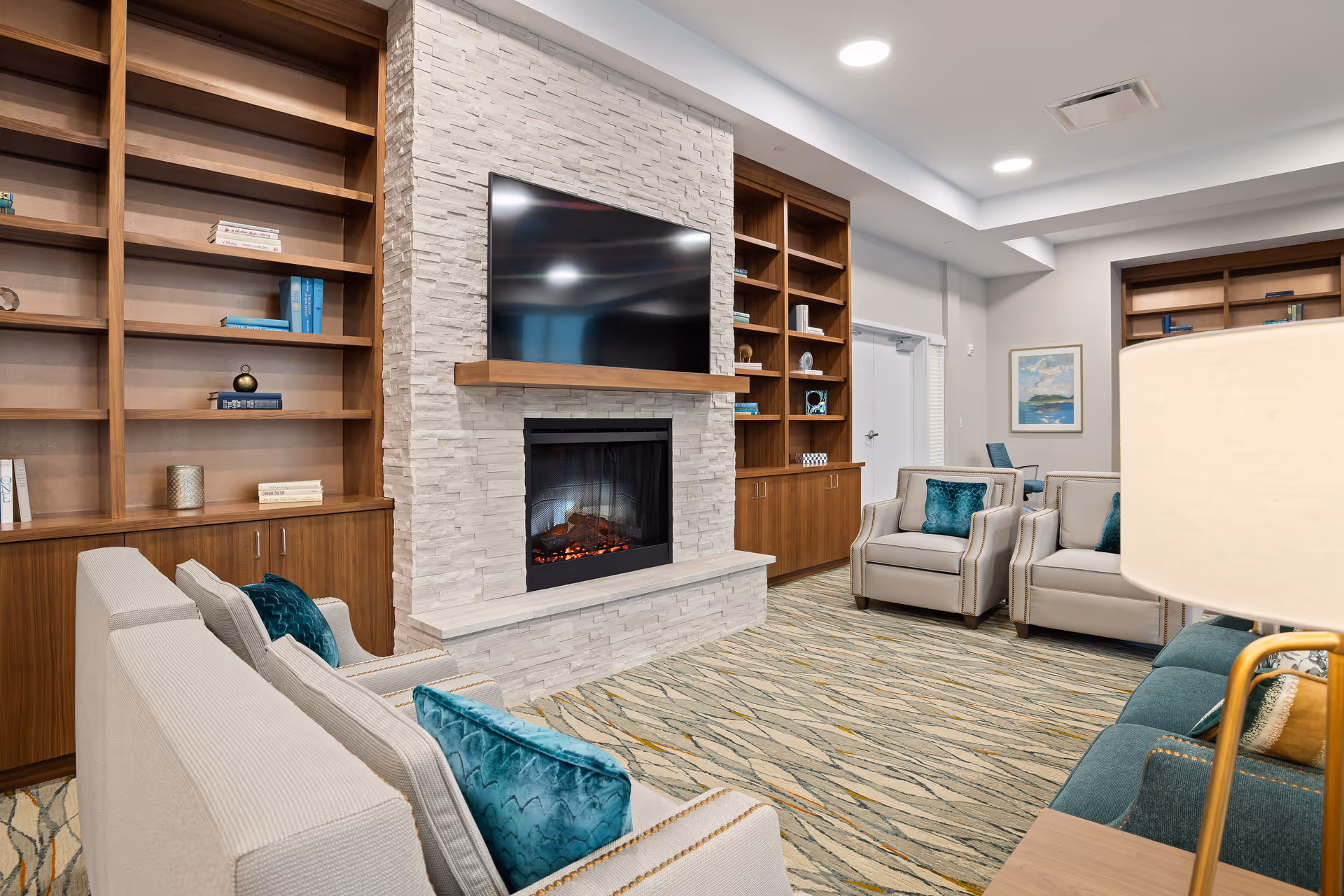 A cozy living room area with beige armchairs and a teal sofa adorned with teal pillows. The room features a modern stone fireplace with a flat-screen TV mounted above it. Surrounding the fireplace are wooden built-in shelves with decorative items and books. The carpet has a patterned design, and the walls are painted white with recessed lighting in the ceiling.