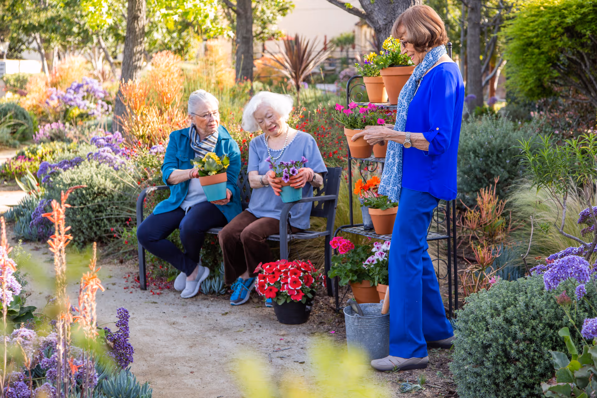 Three elderly women enjoying a garden setting at Hollenbeck Palms. Two women are sitting on a bench holding potted flowers, while the third woman stands nearby arranging more potted plants on a metal plant stand. The garden is lush with colorful flowers and greenery.