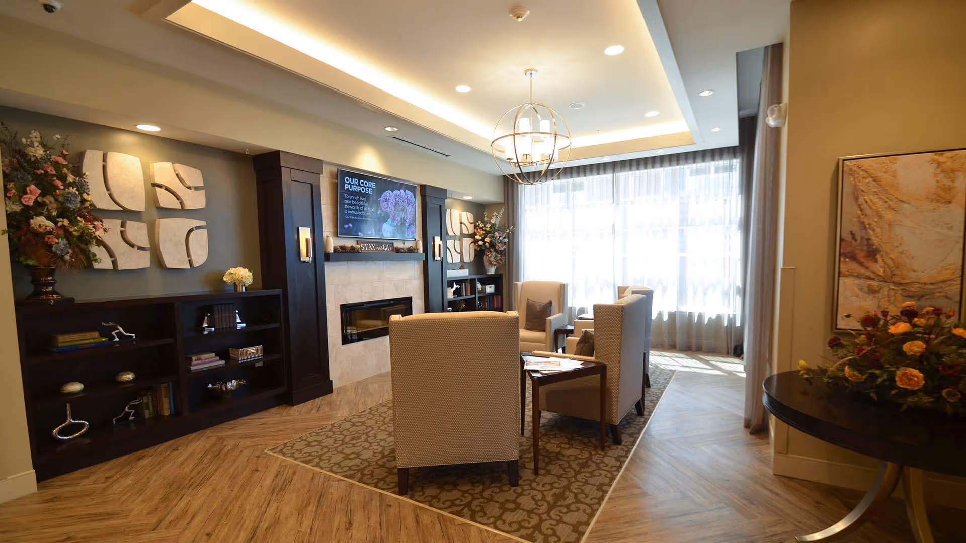 A cozy living room area with four beige armchairs arranged around a small wooden table on a patterned rug. The room features a modern fireplace with a TV mounted above it displaying a message about the facility's core purpose. Decorative wall art and flower arrangements adorn the walls and shelves. Large windows with sheer curtains allow natural light to fill the space, and a round table with a floral arrangement is visible on the right.