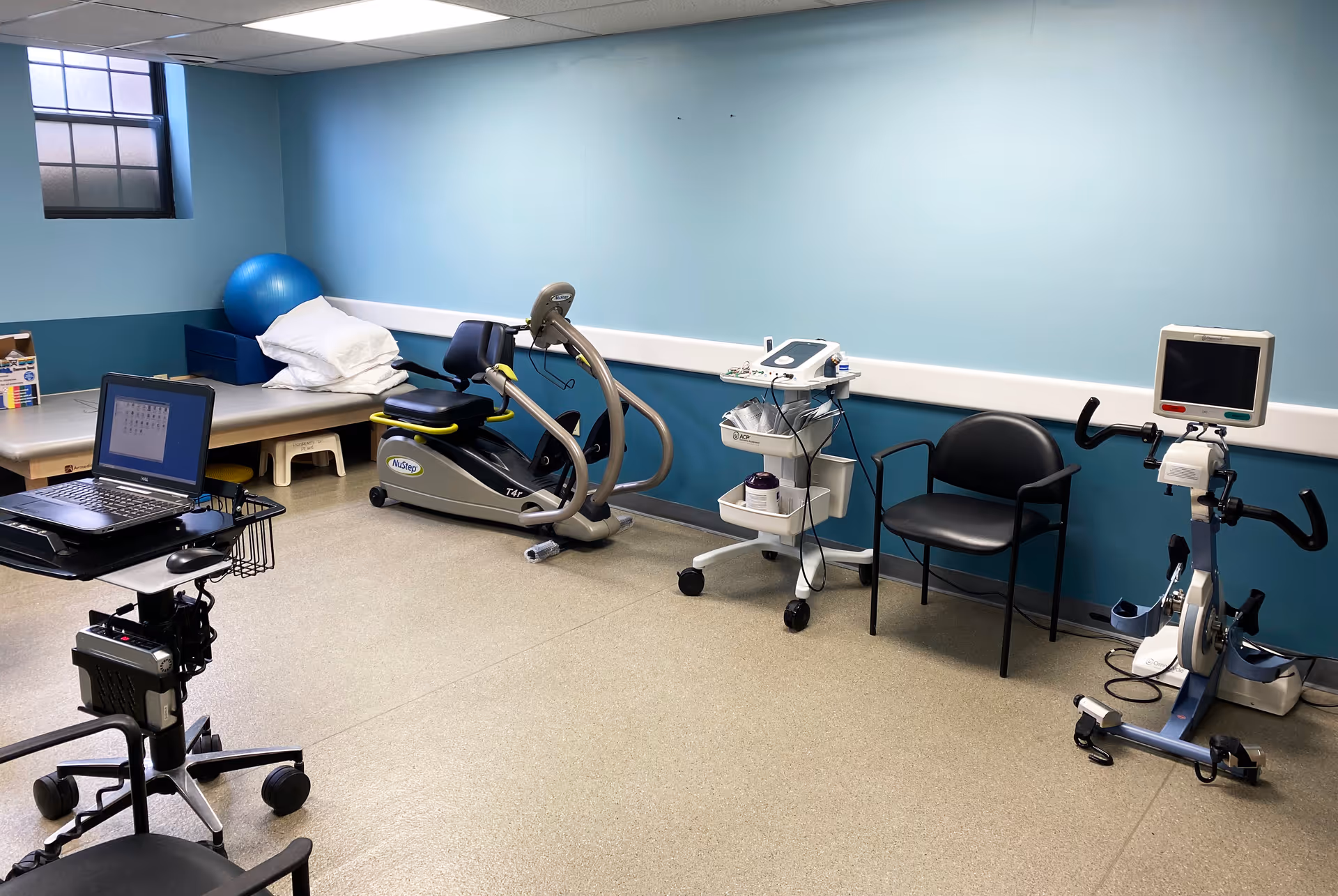 A rehabilitation or physical therapy room with exercise equipment including a recumbent bike, a stationary bike with a monitor, a therapy table with pillows and a blue exercise ball, a black chair, and a medical cart with equipment. The walls are painted blue and there is a small window near the ceiling.
