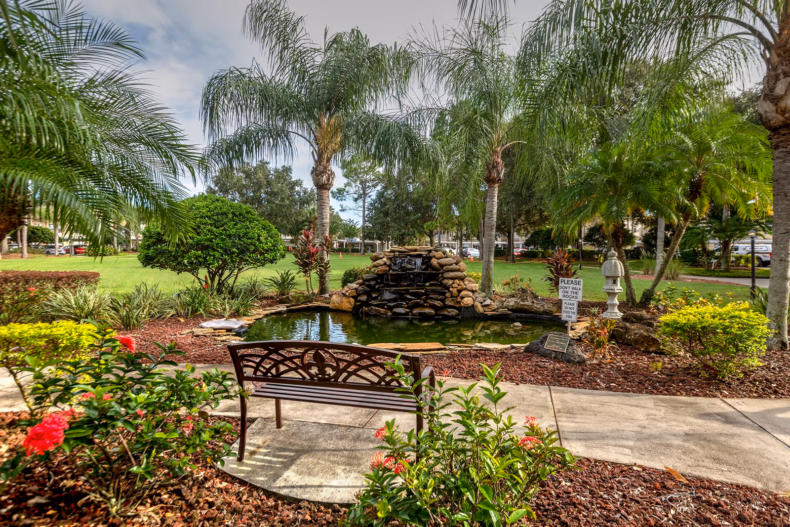 A peaceful outdoor garden area with a small pond and rock waterfall surrounded by palm trees and various plants. A decorative metal bench faces the pond, and a sign near the water reads 'Please don't walk on the rocks. Please do not feed the fish.'