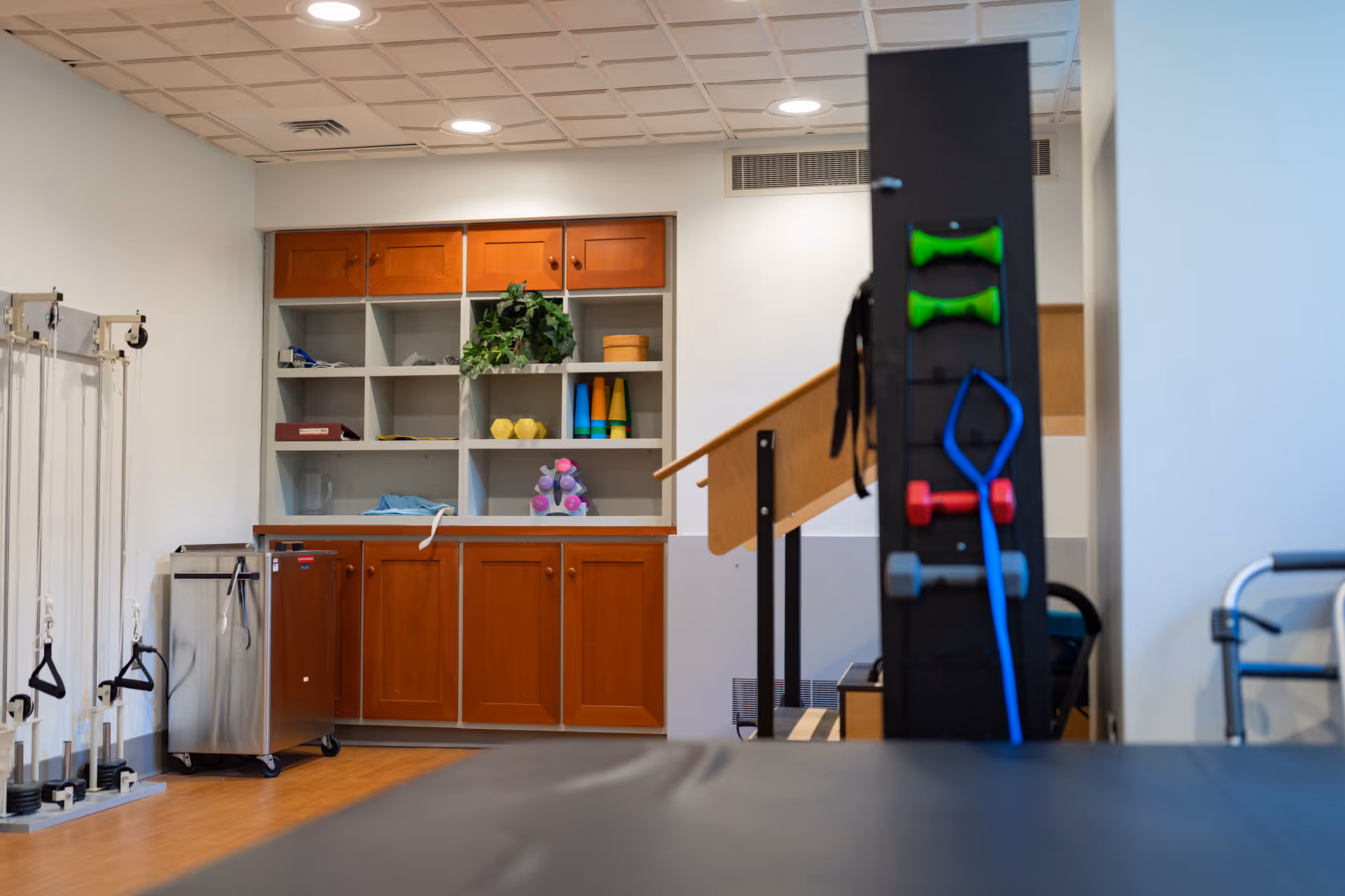 Interior view of a physical therapy or rehabilitation room with wooden cabinets and shelves holding various exercise equipment and supplies. There are resistance bands, small dumbbells, and other therapy tools visible, along with a handrail and exercise machines.