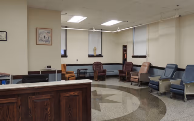 A spacious institutional lounge with a reception desk in the foreground and multiple recliner chairs arranged along the back wall on a patterned terrazzo floor.