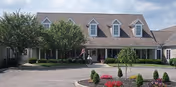 Front exterior of a senior living community building with a circular driveway, landscaped flowerbeds, trees, and a covered entrance displaying an American flag.