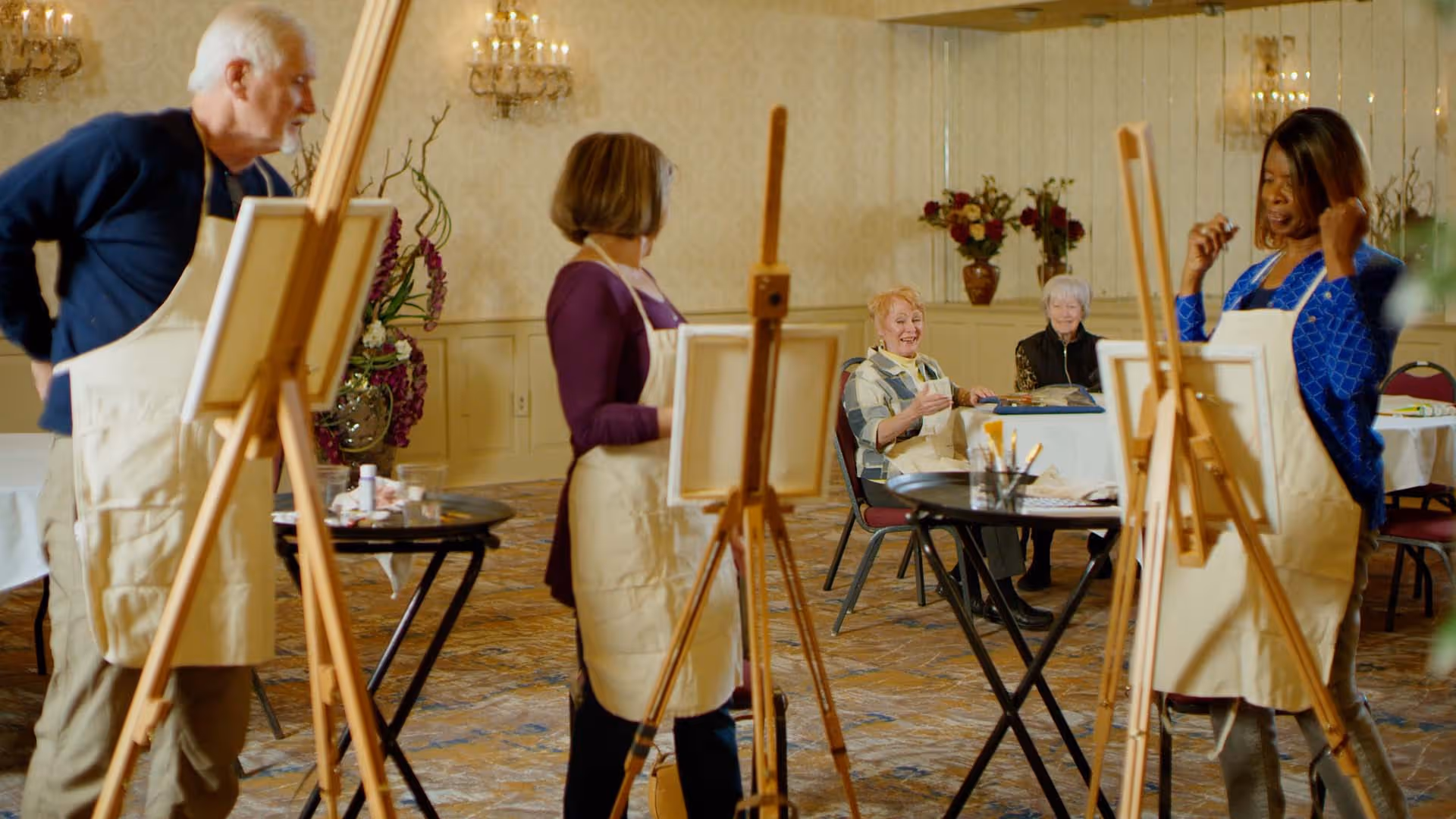 A group of elderly people participating in a painting activity in a well-lit room with chandeliers and floral decorations. Three individuals are standing at easels wearing aprons, while two others are seated at a table in the background, smiling and observing.