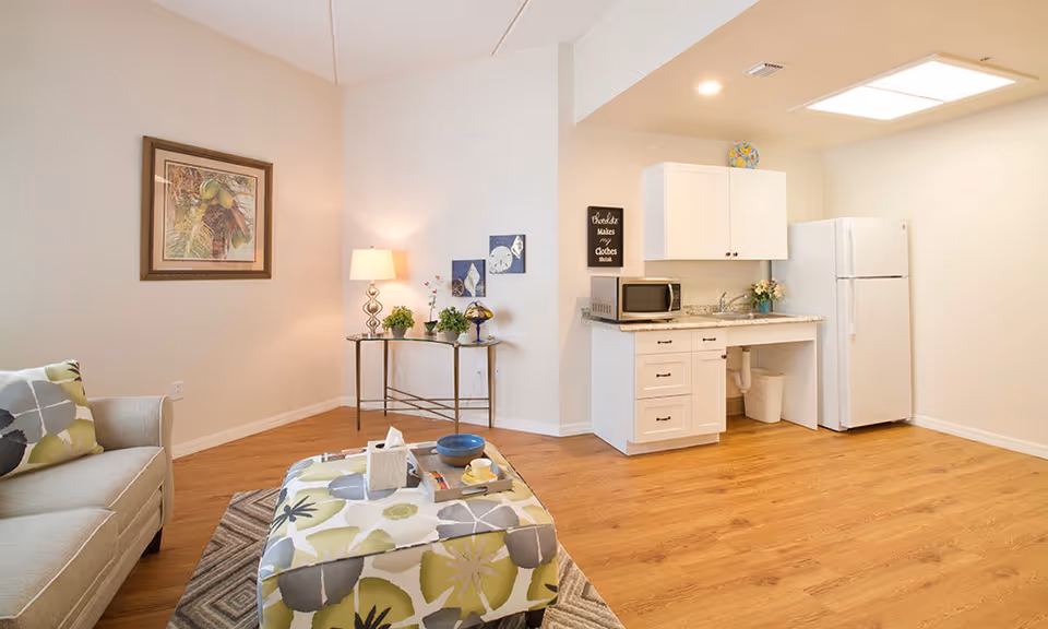 Bright studio-style living area with a sofa and patterned ottoman, a small kitchenette with white cabinets and refrigerator, and decorative table and wall art.