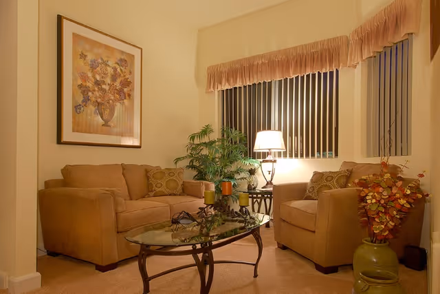 Cozy living room with a beige sofa and armchair, glass coffee table, lamp, plants, and framed artwork.