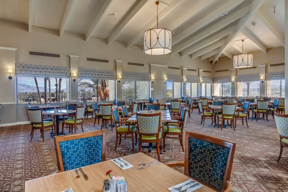 Spacious dining room with multiple wooden tables and chairs featuring green and blue patterned upholstery. Large windows with patterned valances allow natural light to fill the room, and two large pendant lights hang from the vaulted ceiling. The carpet has a geometric pattern, and the room has a bright, welcoming atmosphere.