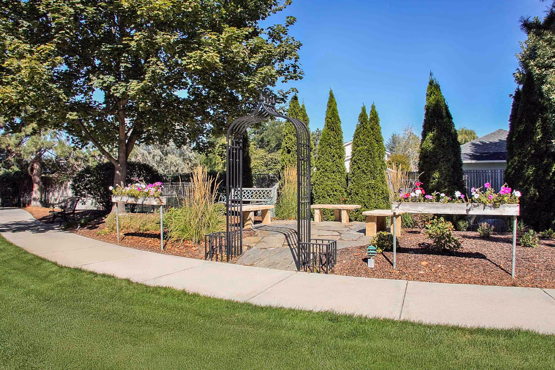 A landscaped outdoor garden with a metal arbor, benches, raised flower planters, trees, and a sidewalk.