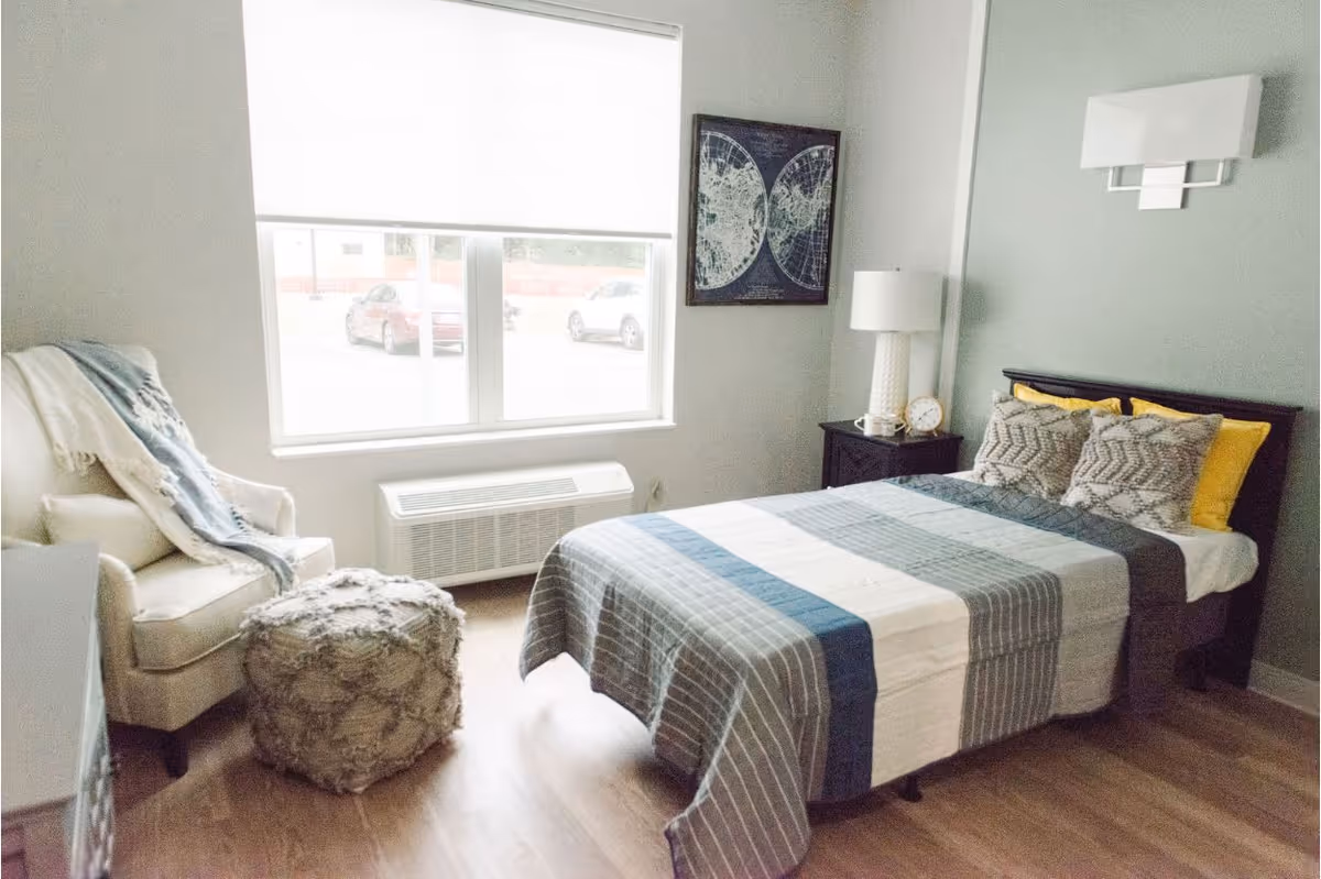 A bright bedroom with a bed covered in a striped quilt and multiple pillows, a nightstand with a lamp and clock, a framed map on the wall, a comfortable armchair with a throw blanket, and a textured ottoman. A large window lets in natural light and shows parked cars outside.