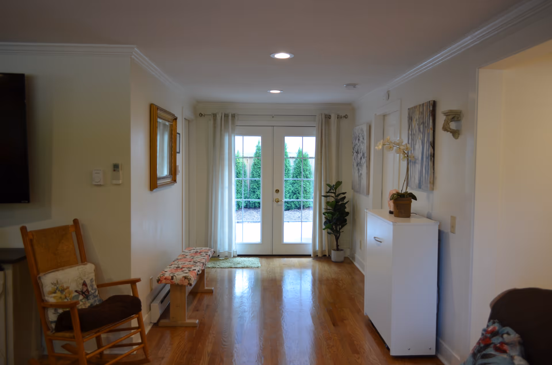 Hallway-like living area with hardwood floors, a rocking chair and bench, a white cabinet and potted plants facing glass French doors.