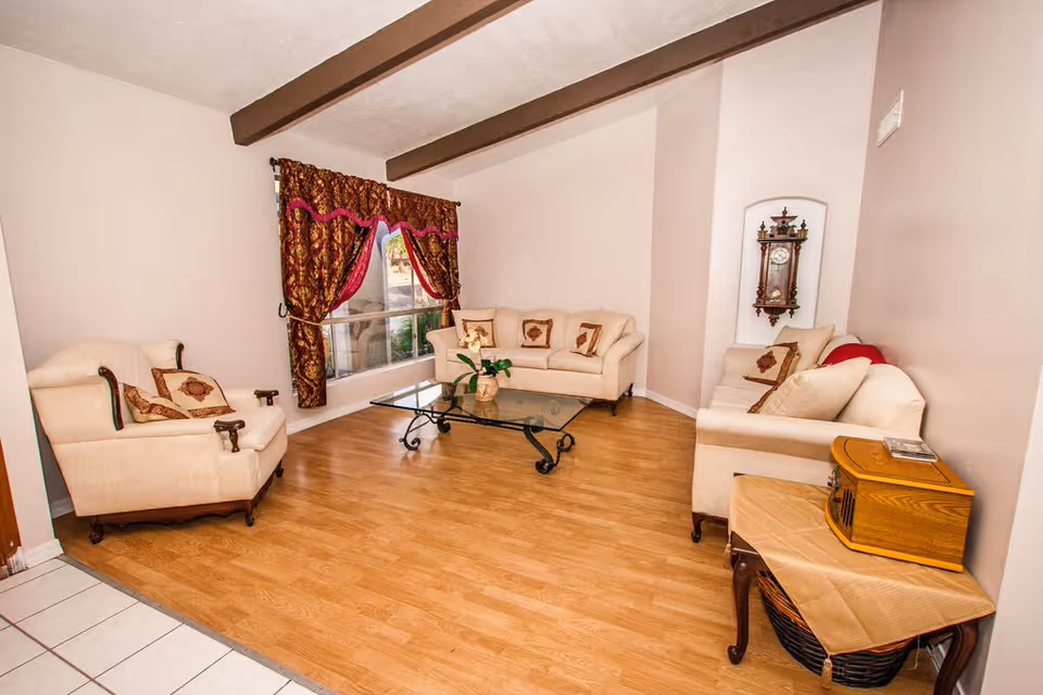 A cozy living room with light beige walls and wooden floor. The room features three cream-colored upholstered sofas with decorative pillows, a glass coffee table with a plant centerpiece, a large window with ornate red and gold curtains, exposed wooden ceiling beams, and a vintage wall clock. There is also a small wooden table with a basket underneath and a wooden box on top, covered with a beige cloth.