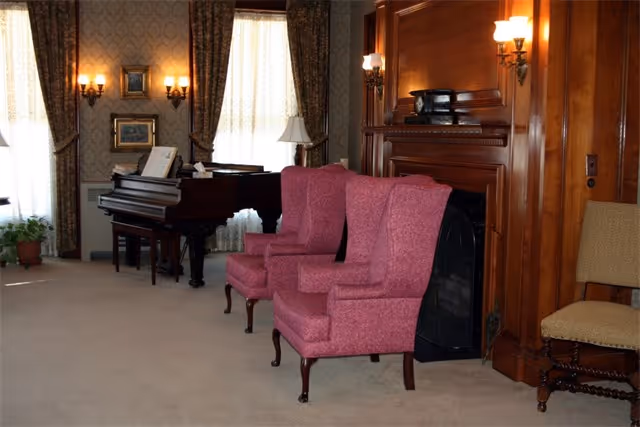 A cozy living room with two pink upholstered armchairs placed in front of a wooden fireplace. To the left, there is a grand piano with sheet music on it, and the room is decorated with patterned wallpaper, framed pictures, wall sconces, and long curtains covering the windows. A small potted plant is visible near the window.