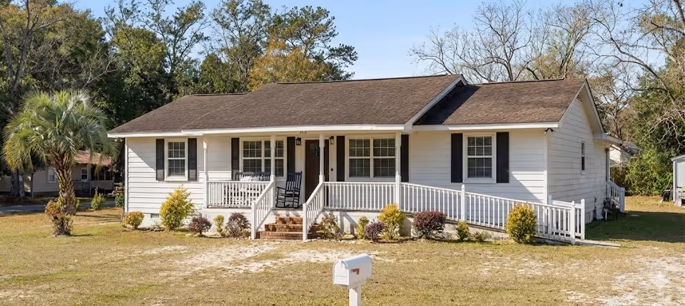 Single-story white house with a front porch, wheelchair-access ramp, and a mailbox on a grassy yard.