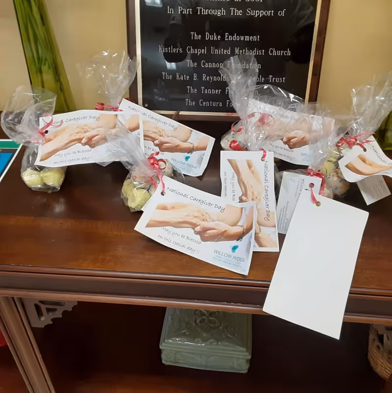 A wooden table with several small gift bags wrapped in clear plastic and tied with red ribbons. Each bag contains yellow and multicolored items and has a card attached that reads 'National Caregiver Day' and 'May you be blessed on this special day!!' with an image of two hands holding each other. Behind the table is a plaque with various names and organizations listed.