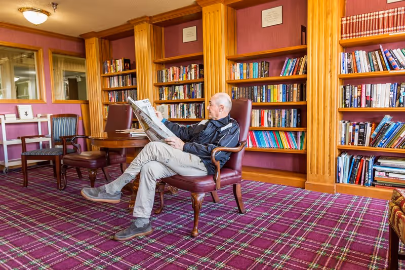 An elderly man sitting in a leather armchair reading a newspaper in a cozy library room with wooden bookshelves filled with books and a purple plaid carpet.