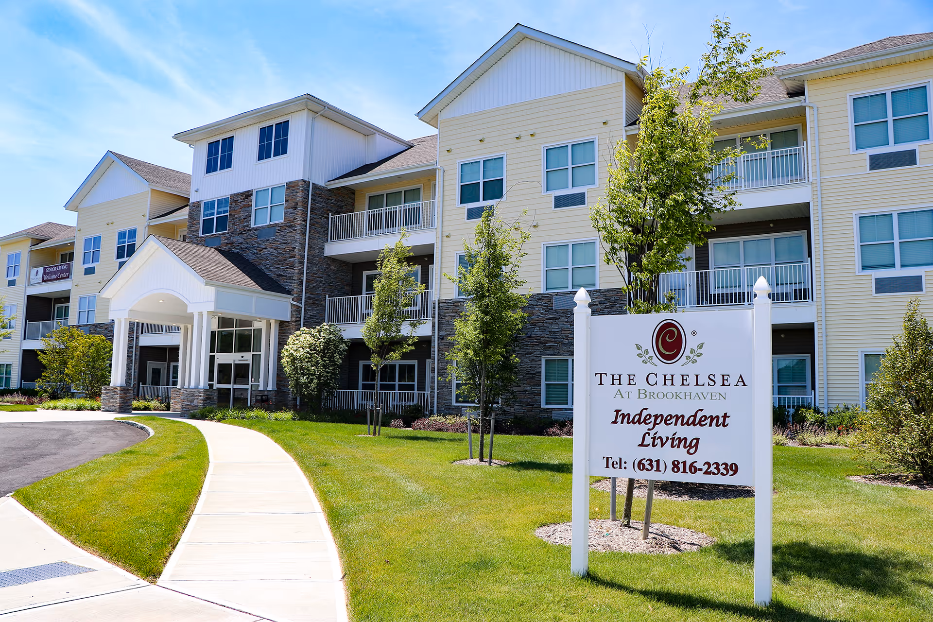 Exterior view of The Chelsea at Brookhaven senior living facility on a sunny day, showing a multi-story building with balconies, a covered entrance, well-maintained lawn, trees, and a sign in the foreground that reads 'The Chelsea at Brookhaven Independent Living Tel: (631) 816-2339'.