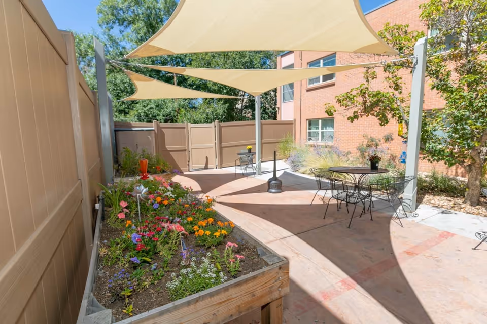 Outdoor patio area with shade sails overhead, metal tables and chairs, and a raised garden bed filled with colorful flowers. The area is enclosed by a beige fence and adjacent to a brick building with windows. Trees and greenery are visible in the background.