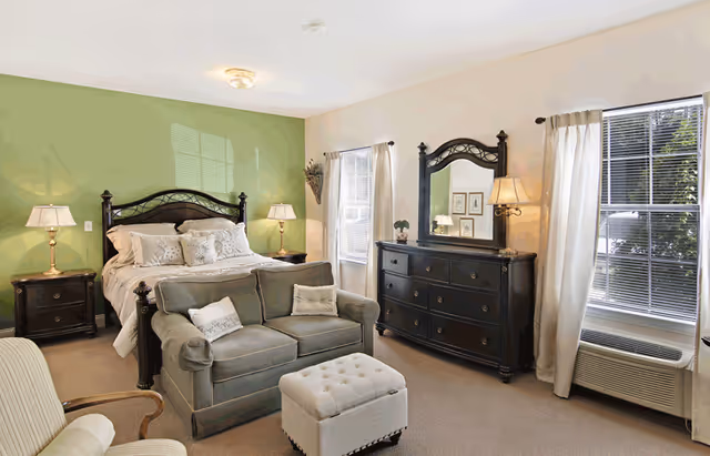 A well-lit bedroom with a large bed featuring a dark wooden headboard against a green accent wall. The bed is flanked by two nightstands with lamps. In front of the bed is a small green loveseat with two pillows and a light-colored tufted ottoman. To the right, there is a dark wooden dresser with a large mirror and a wall-mounted lamp. Two windows with white curtains allow natural light into the room. A beige armchair is partially visible in the foreground.