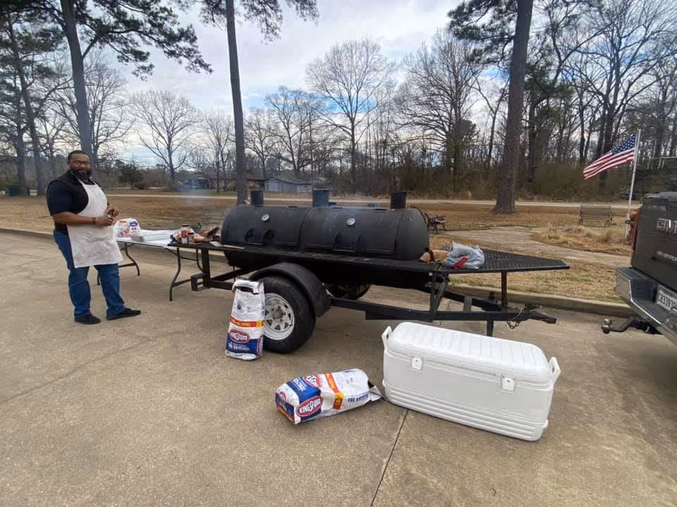 A man wearing an apron stands next to a large black barbecue smoker on a trailer in an outdoor parking area. There are bags of charcoal and a white cooler on the ground nearby. Trees and a flagpole with an American flag are visible in the background.