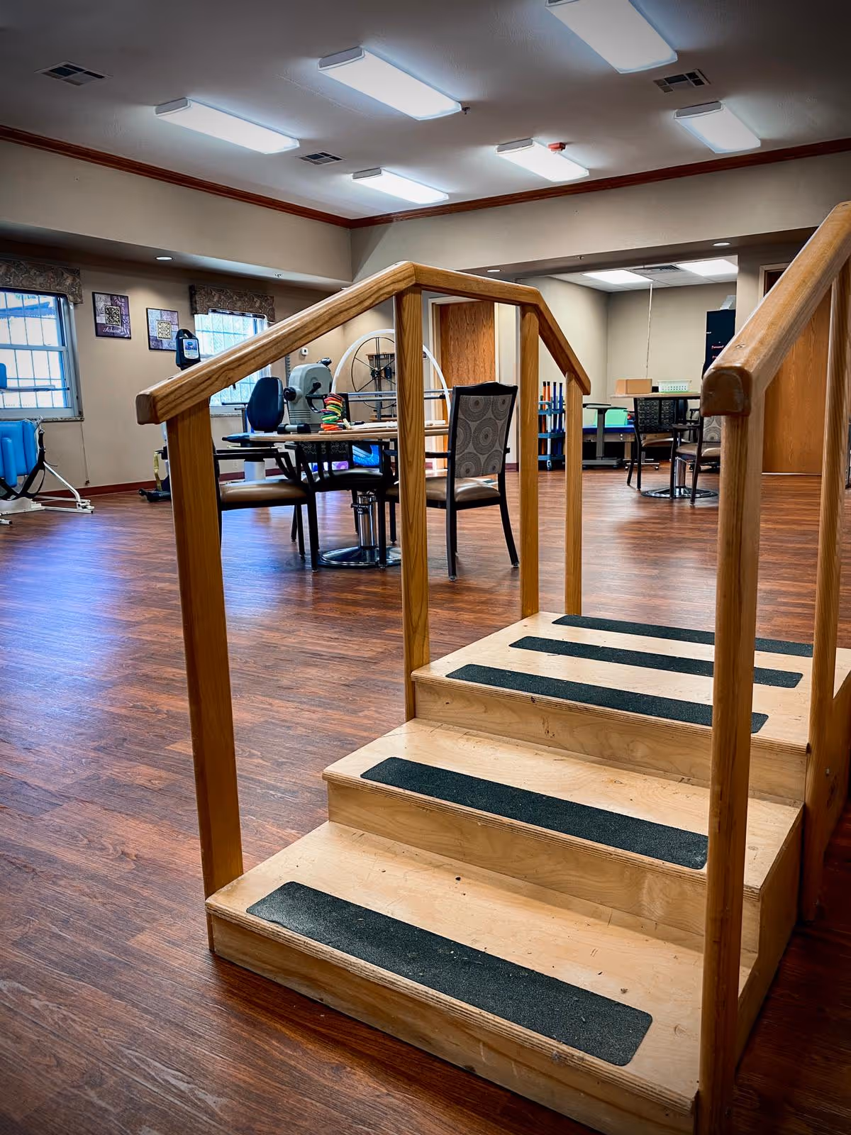 Interior view of a rehabilitation or therapy room with wooden flooring, a small wooden staircase with handrails and anti-slip strips, tables and chairs, exercise equipment, and windows with blinds.