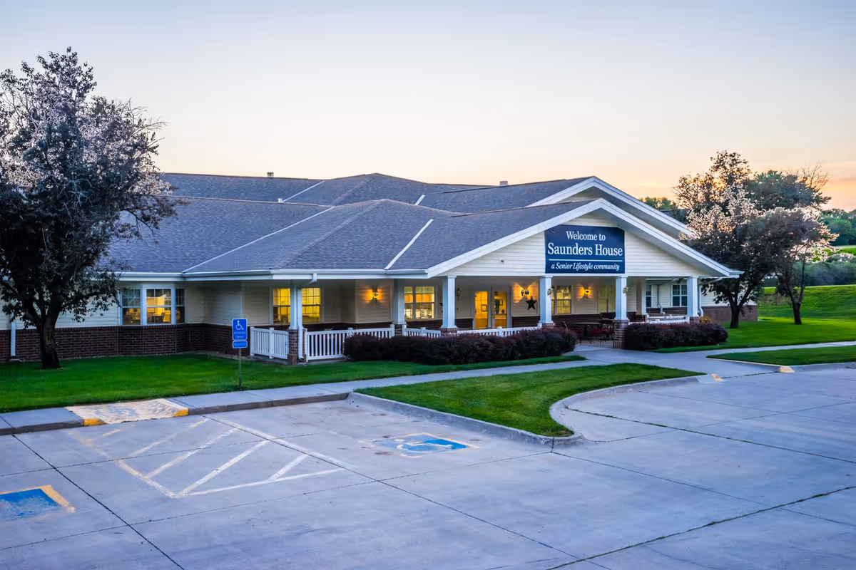 Front exterior of Saunders House senior living facility with a covered porch, illuminated windows, lawn and parking area at dusk.