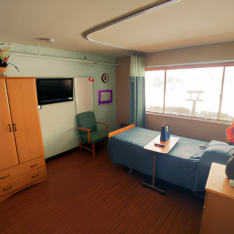 A small, tidy room with a single bed covered in blue bedding, a wooden wardrobe, a green armchair, a wall-mounted TV, a window with curtains, and a small table on wheels placed over the bed.