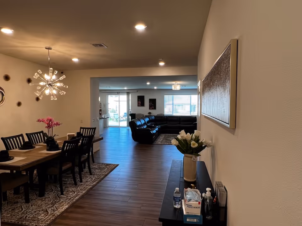 Interior view of a senior living facility showing a dining area with a wooden table and six chairs on a patterned rug, a modern chandelier above the table, and a living room with a large black sectional sofa in the background. There is a console table with a vase of white flowers, hand sanitizer, face masks, and a water bottle along the right wall. The room has wooden flooring and neutral-colored walls with some wall art.