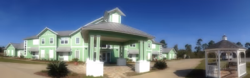 Exterior view of a large green two-story senior living facility with white trim and a covered entrance. The building is surrounded by a paved driveway and landscaped with small bushes. To the right, there is a white gazebo on a paved area under a clear blue sky.