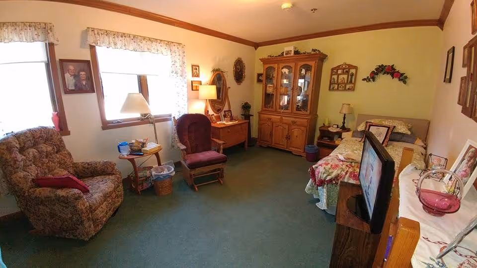 A cozy bedroom with a single bed covered in floral bedding, a wooden cabinet with glass doors, a wooden desk with a lamp and framed photos, a cushioned armchair, a rocking chair, and a TV on a stand. The room has green carpet, light-colored walls with framed pictures, and two windows with floral curtains letting in natural light.