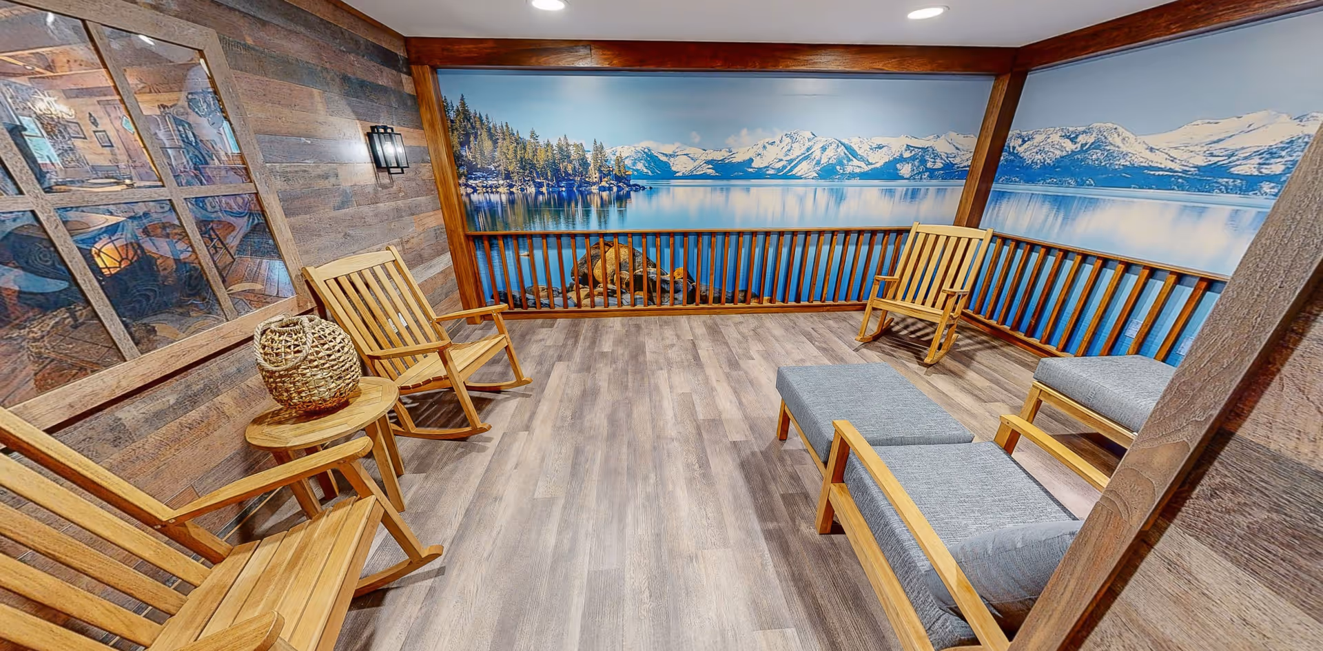 A cozy indoor seating area with wooden rocking chairs and cushioned benches arranged on a wood-patterned floor. The walls are decorated with wood paneling and a large mural depicting a serene lake with snow-capped mountains and pine trees. A small wooden side table with a woven basket is placed next to one of the rocking chairs.
